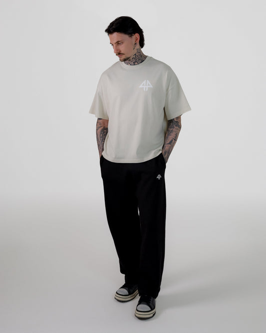 A man modeling a light beige short-sleeve t-shirt with a small white graphic on the chest, paired with black sweatpants and black-and-white sneakers, standing against a light gray studio background.