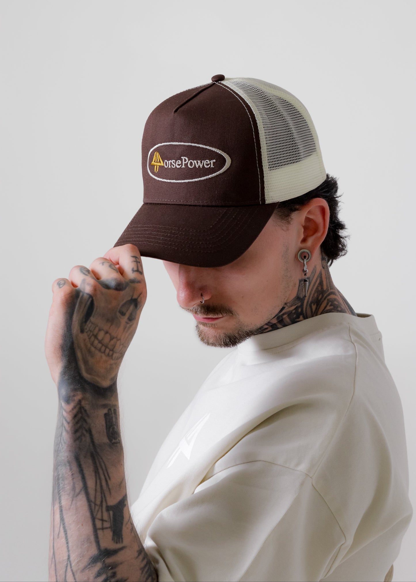 A man wearing a brown and cream trucker hat with a 'HorsePower' logo, holding the brim of the hat. The background is a clean, neutral light grey studio backdrop.