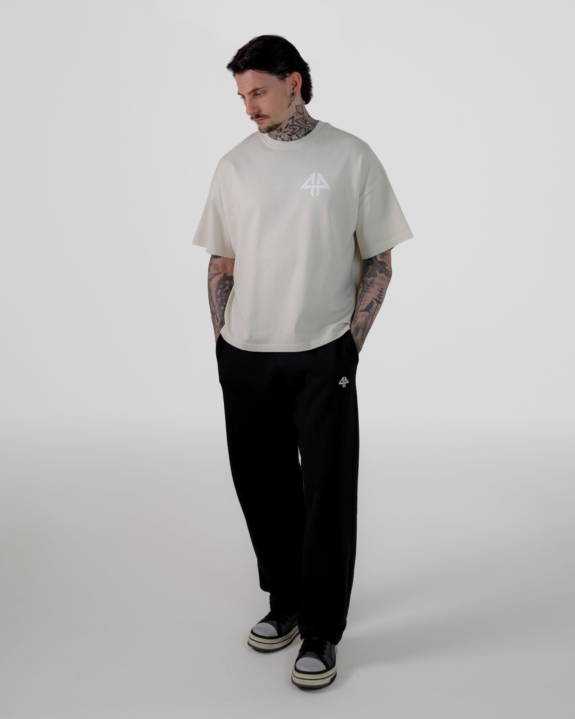 A man modeling a light beige short-sleeve t-shirt with a small white graphic on the chest, paired with black sweatpants and black-and-white sneakers, standing against a light gray studio background.