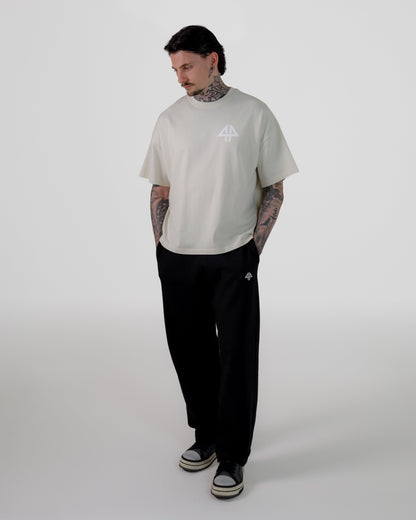 A man modeling a light beige short-sleeve t-shirt with a small white graphic on the chest, paired with black sweatpants and black-and-white sneakers, standing against a light gray studio background.