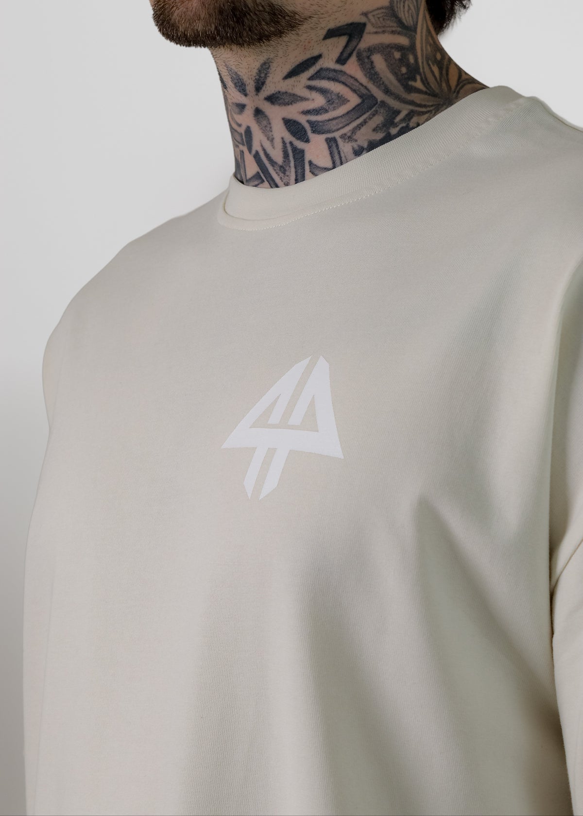 A close-up of a man wearing a light beige crewneck sweatshirt with a white logo on the chest, set against a plain light grey background.