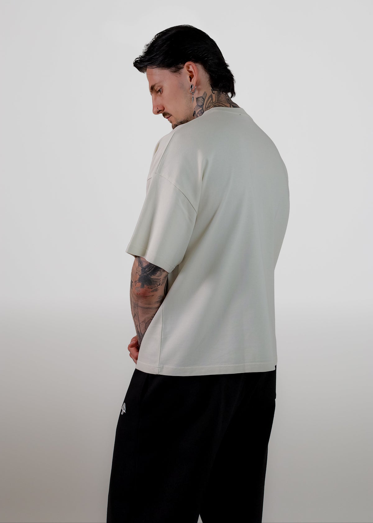 A man with tattoos is shown from a side-back angle wearing an oversized off-white t-shirt and black sweatpants against a plain light gray background.