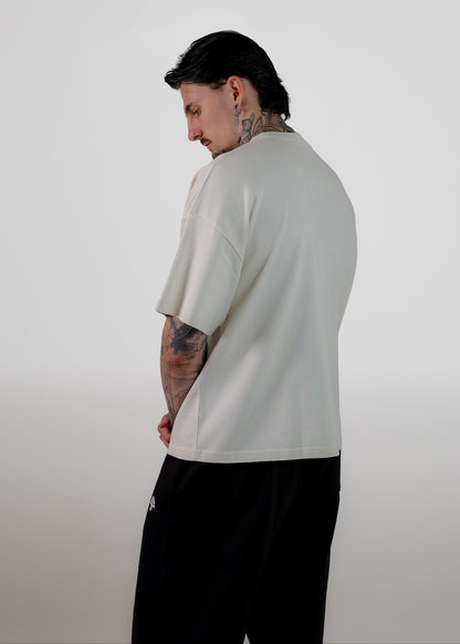 A man with tattoos is shown from a side-back angle wearing an oversized off-white t-shirt and black sweatpants against a plain light gray background.