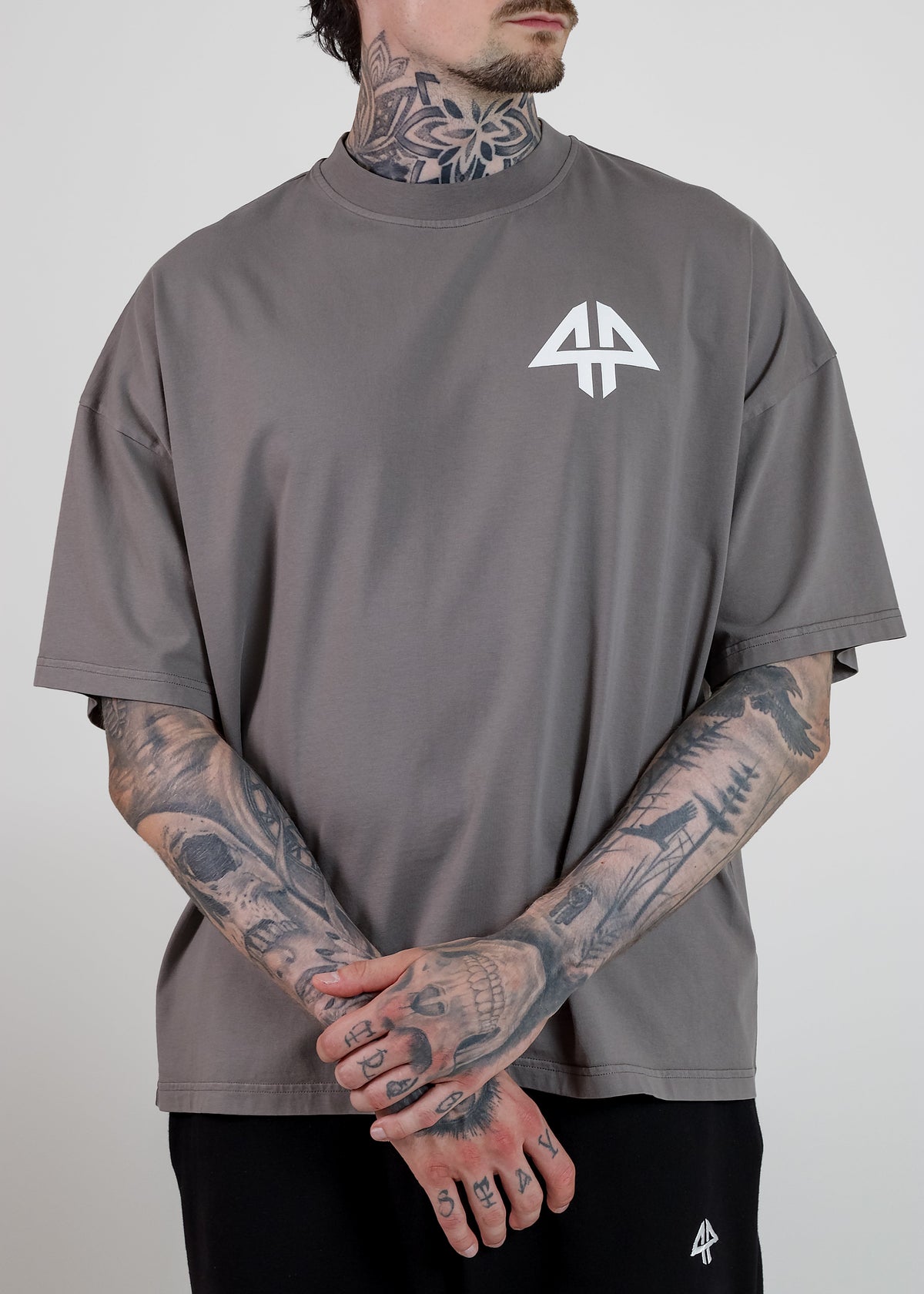 A man wearing an oversized taupe grey t-shirt with a white geometric logo on the left chest, paired with black sweatpants. The photo is cropped from the chin down against a neutral light grey background.