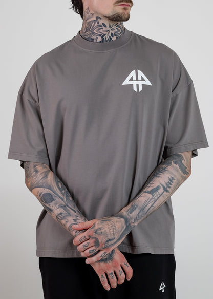 A man wearing an oversized taupe grey t-shirt with a white geometric logo on the left chest, paired with black sweatpants. The photo is cropped from the chin down against a neutral light grey background.