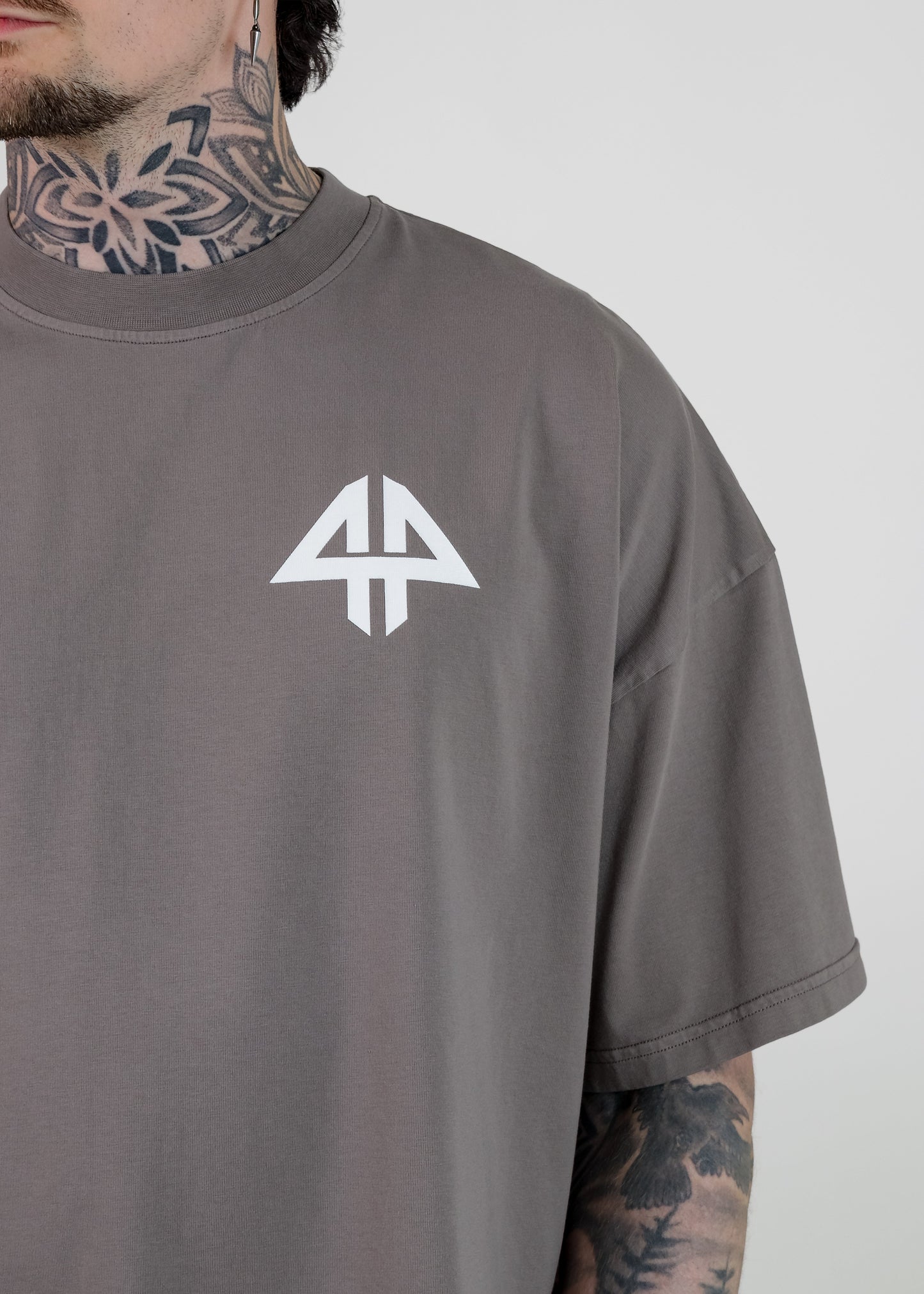 A close-up of a man wearing a dark gray t-shirt with a white geometric logo on the chest, showing his tattooed neck and arm against a light gray background.