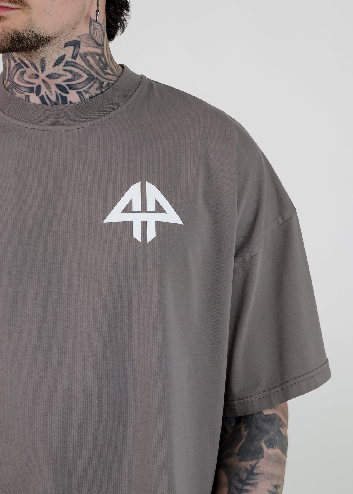 A close-up of a man wearing a dark gray t-shirt with a white geometric logo on the chest, showing his tattooed neck and arm against a light gray background.