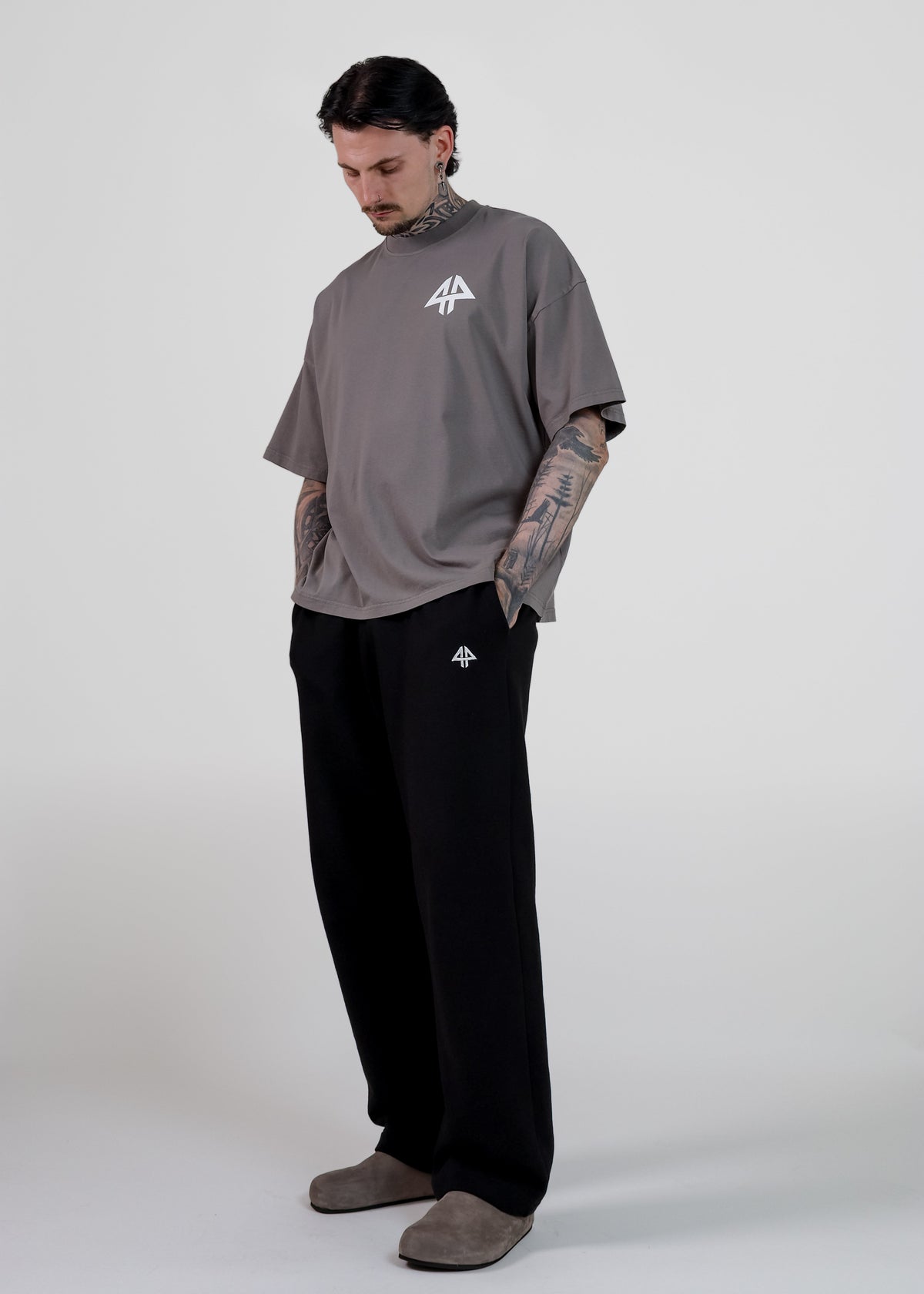 A man modelling a loose-fit taupe grey t-shirt and black sweatpants with small white logo details, standing against a light grey studio background.