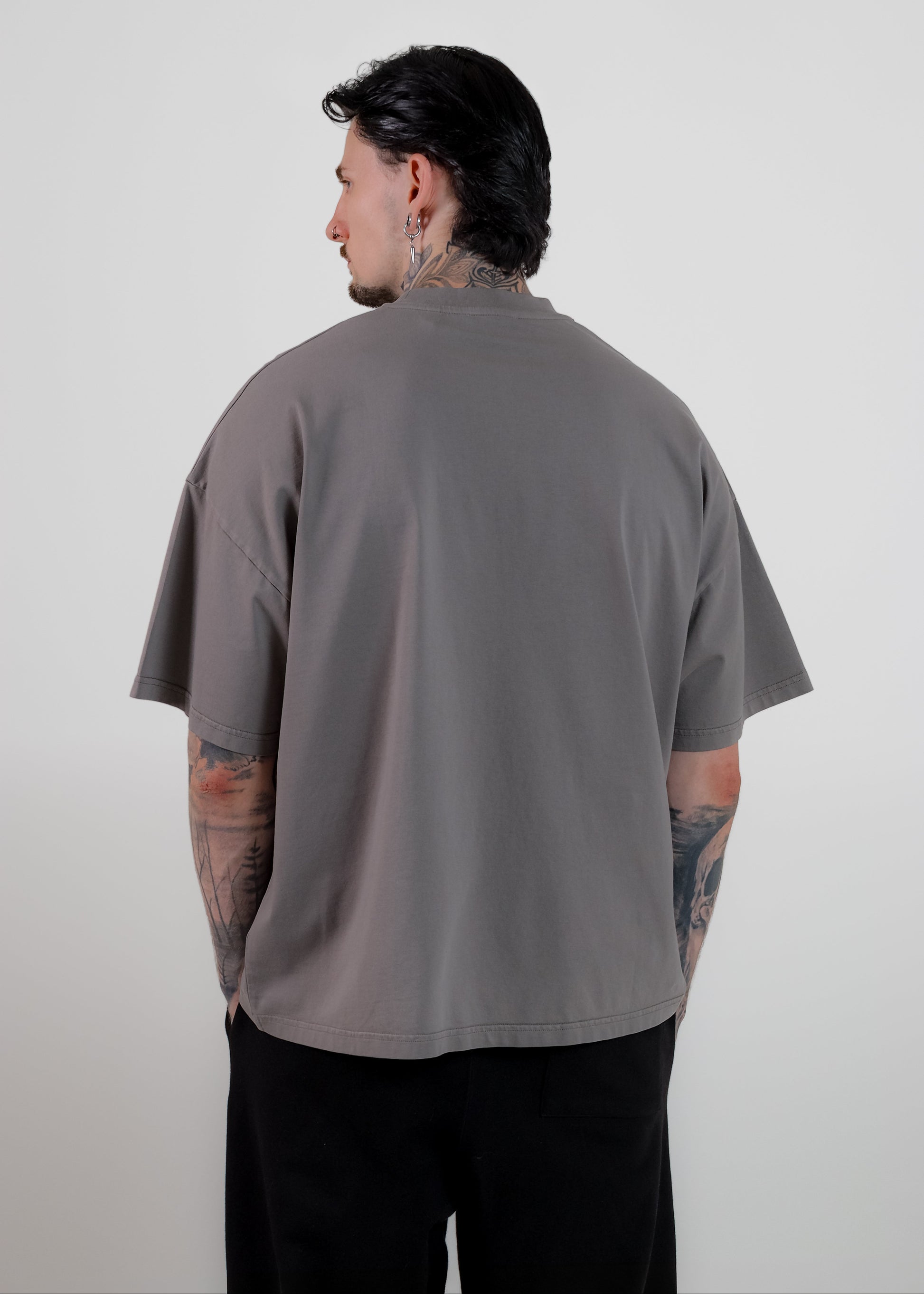 A man with tattoos is shown from the back wearing an oversized, short-sleeve grey t-shirt and black pants against a plain light grey background.