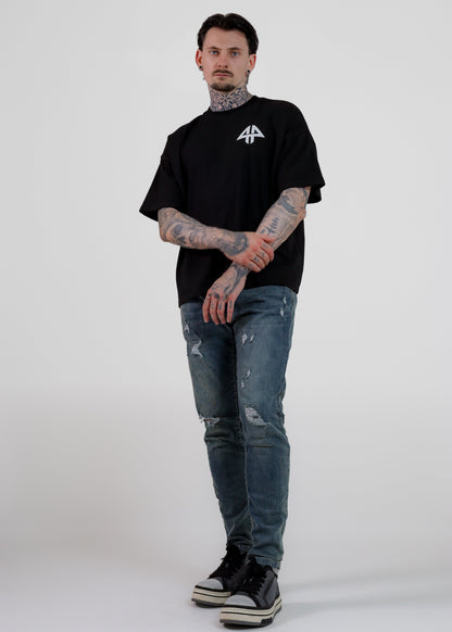 A man with tattoos stands against a light grey studio background wearing a black short-sleeve t-shirt with a white graphic logo on the chest, distressed blue jeans, and black sneakers.