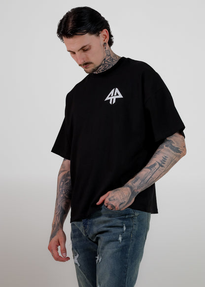A man with tattoos wearing an oversized black t-shirt with a white geometric logo on the chest, paired with distressed blue jeans, standing against a light grey background.