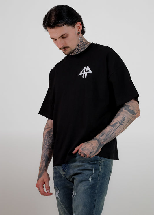 A man with tattoos wearing an oversized black t-shirt with a white geometric logo on the chest, paired with distressed blue jeans, standing against a light grey background.
