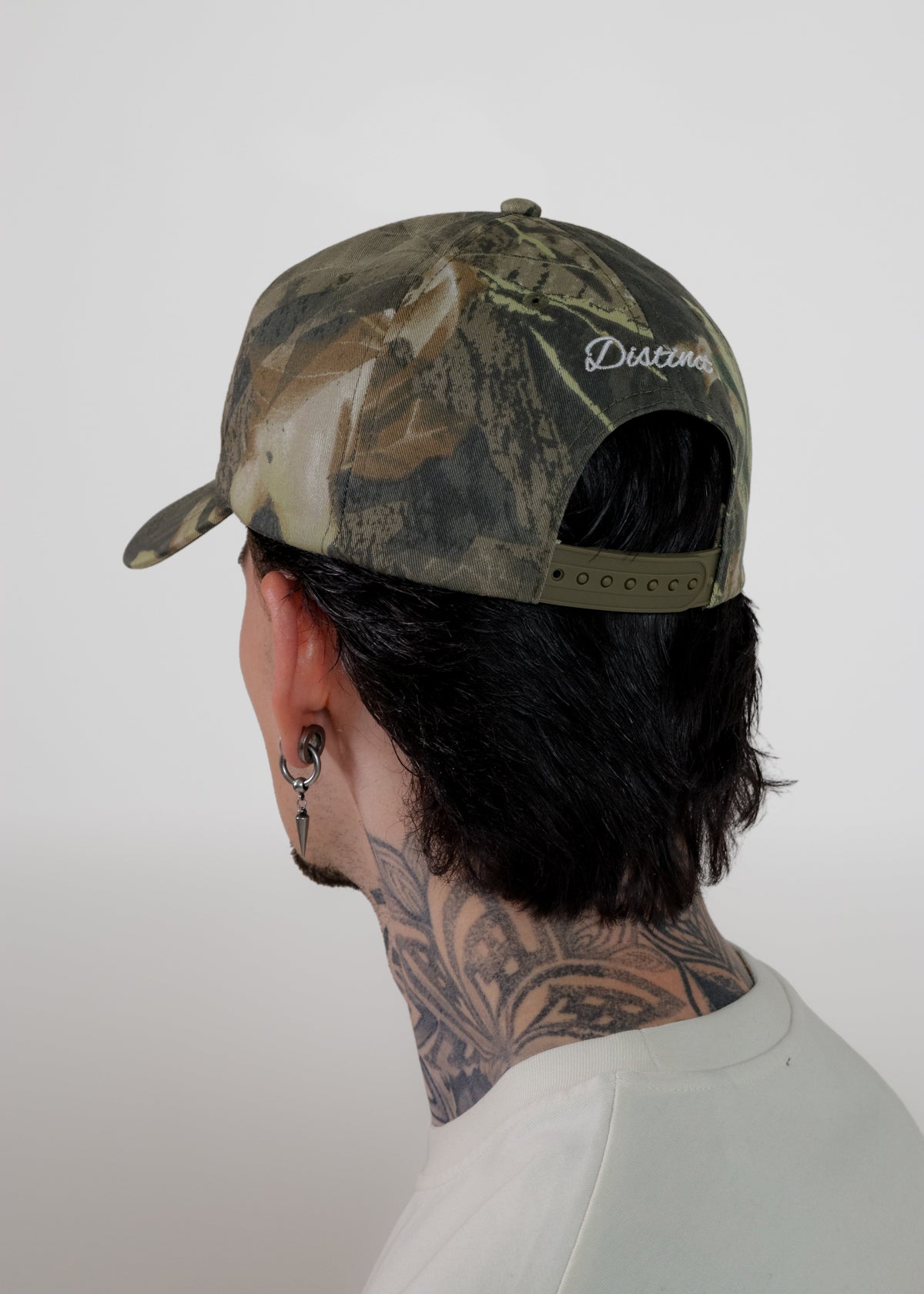 A man shown from the back wearing a camouflage baseball cap with a visible 'Distinct' logo on the back, set against a plain light grey background.