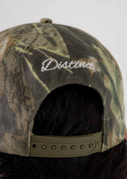 Close-up back view of a camouflage baseball cap with 'Distinct' embroidered in white script on the crown, shown on a person with dark hair against a light gray background.