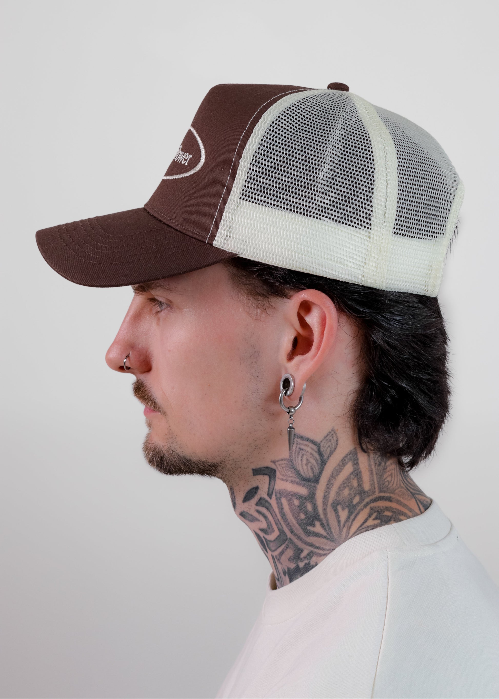 A man in profile wearing a brown and white trucker hat with a mesh back, against a plain light grey background.