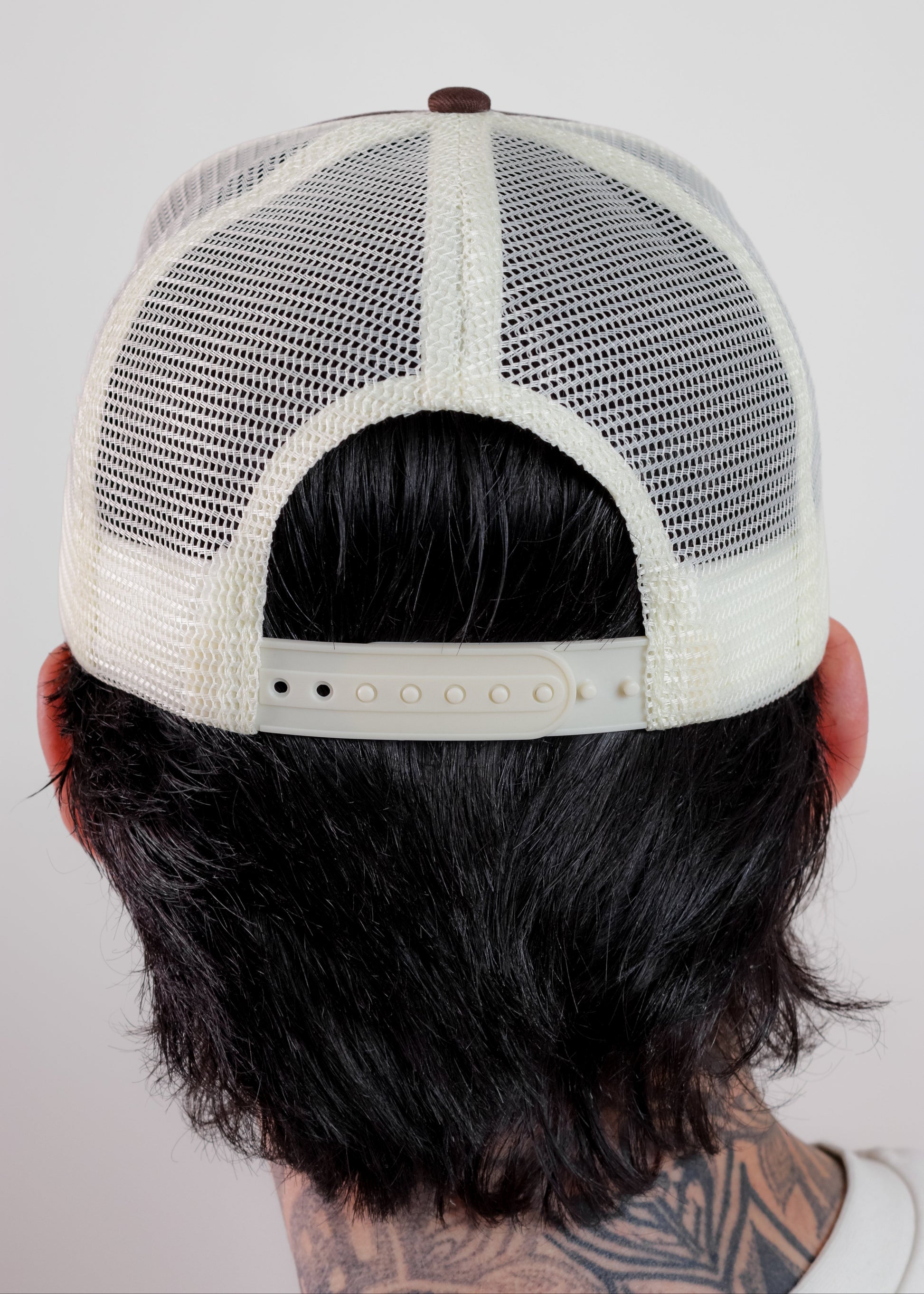 Back view of a person wearing a cream-colored trucker hat with a white mesh back and adjustable strap, set against a plain light gray background.