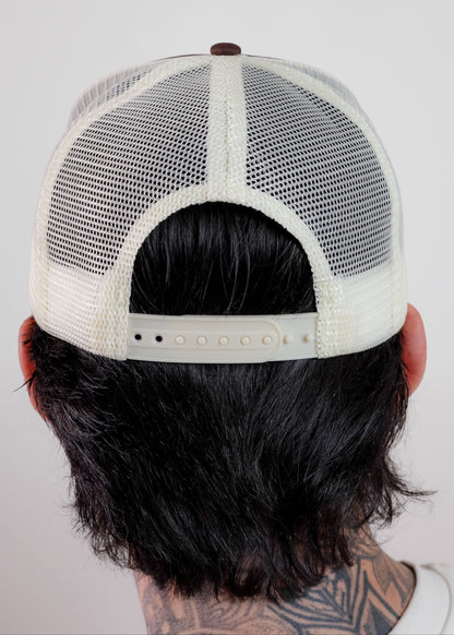 Back view of a person wearing a cream-colored trucker hat with a white mesh back and adjustable strap, set against a plain light gray background.